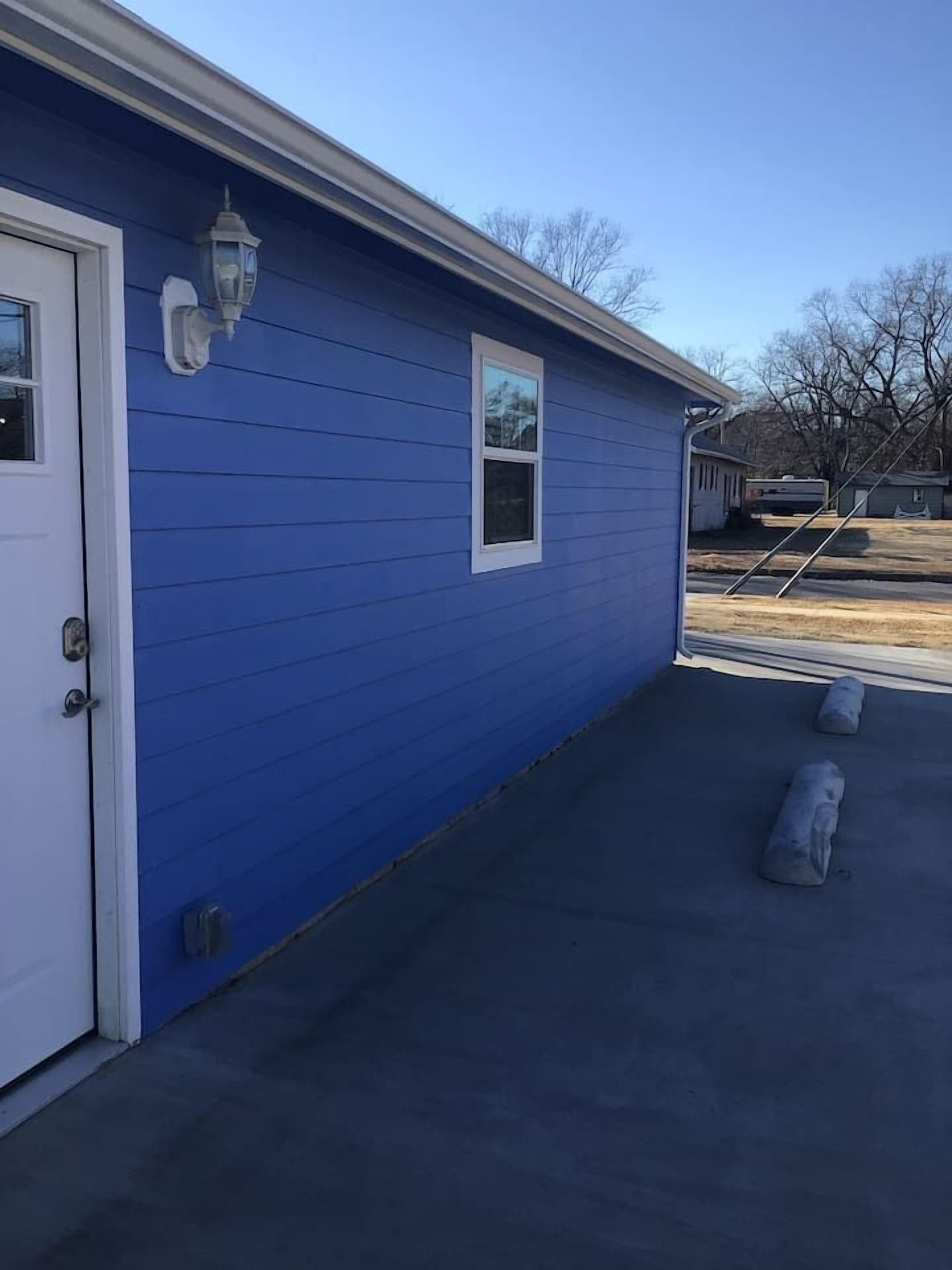 A blue building with a white door and a window, a concrete driveway, and a metal railing in the background.