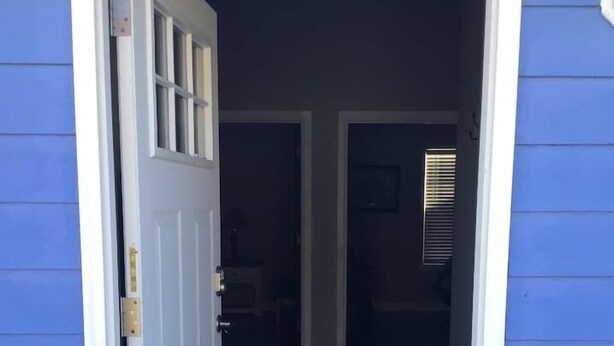 Une porte d’entrée mène à un espace intérieur faiblement éclairé, où l’on aperçoit un banc et le cadre d’une porte.