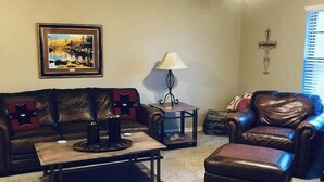 A living room with a sofa, a coffee table, a leather armchair, a lamp, and a ceiling fan. - MJ’S BUNKHOUSE #3-2 BLOCKS TO DOWNTOWN (Pawhuska)
