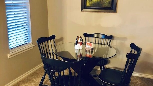 A dining area with a round table and four chairs, a chandelier, a painting on the wall, and a window with blinds. - MJ’S BUNKHOUSE #3-2 BLOCKS TO DOWNTOWN (Pawhuska)
