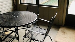 A patio area with a round metal table and four chairs, a window with a beige blind, and a view of a wooden fence. - MJ’S BUNKHOUSE #3-2 BLOCKS TO DOWNTOWN (Pawhuska)