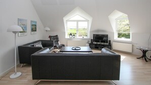 A modern living room with a dark leather sofa, a coffee table, and a TV. There are two windows with white frames and a view of greenery outside. - 3 bedroom gorgeous apartment in Samsø (Samsø)