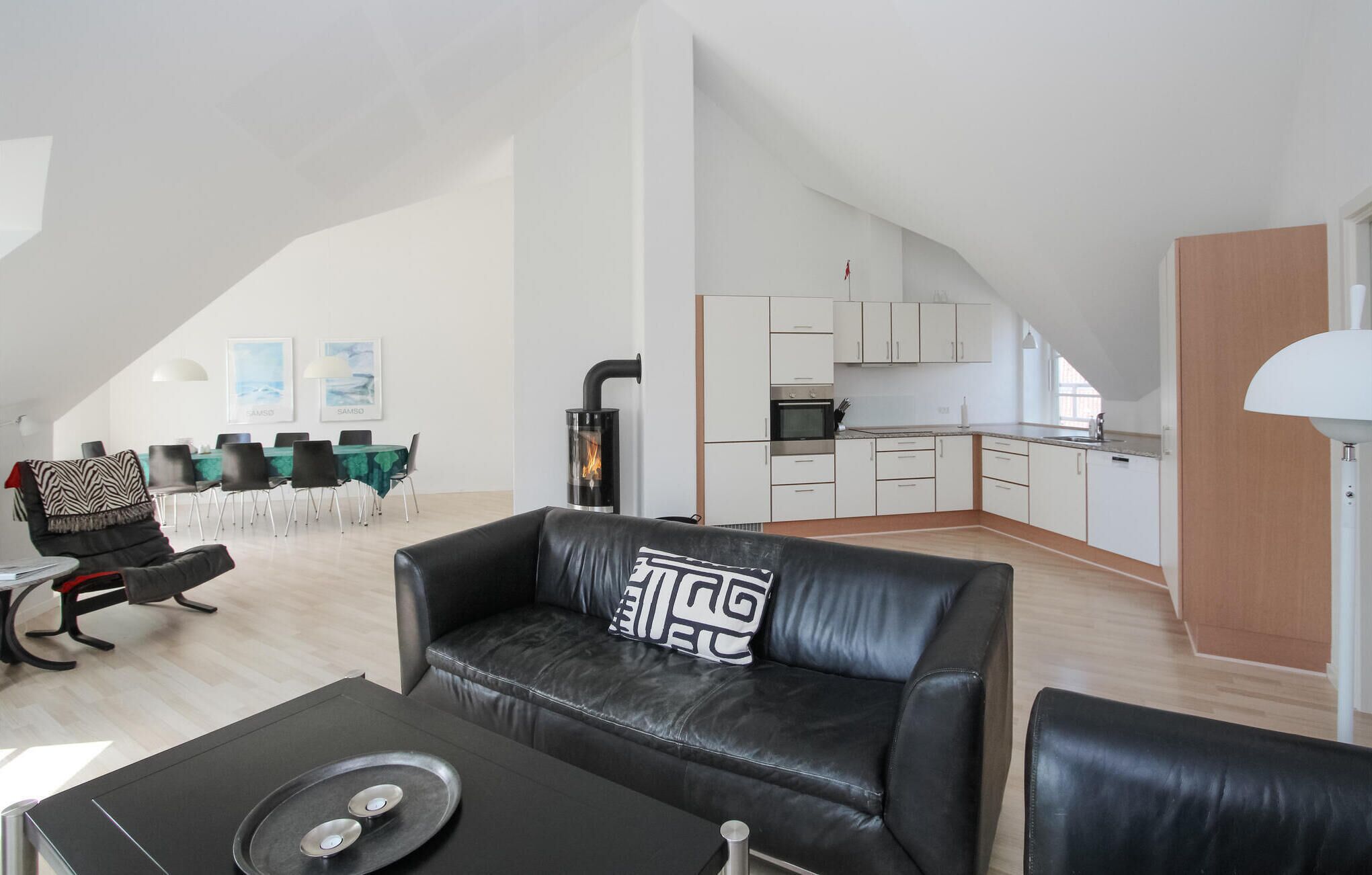 Een modern loft appartement met een hoge plafond, een eetgedeelte met een groene tafel, een zwarte leren bank met een geometrisch kussen, een keuken met witte kasten, en een groot raam.