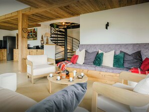 Una sala de estar moderna con un sofá gris, sillones blancos y una mesa de centro de madera. Se ve una escalera con barandilla negra al fondo.