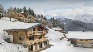 A wooden chalet with a balcony, surrounded by snow-covered trees and mountains.