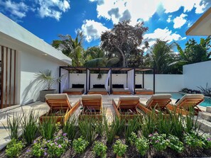 A modern outdoor lounge area with wooden loungers, a covered seating area, and a pool. - Illa Cozumel (Cozumel)