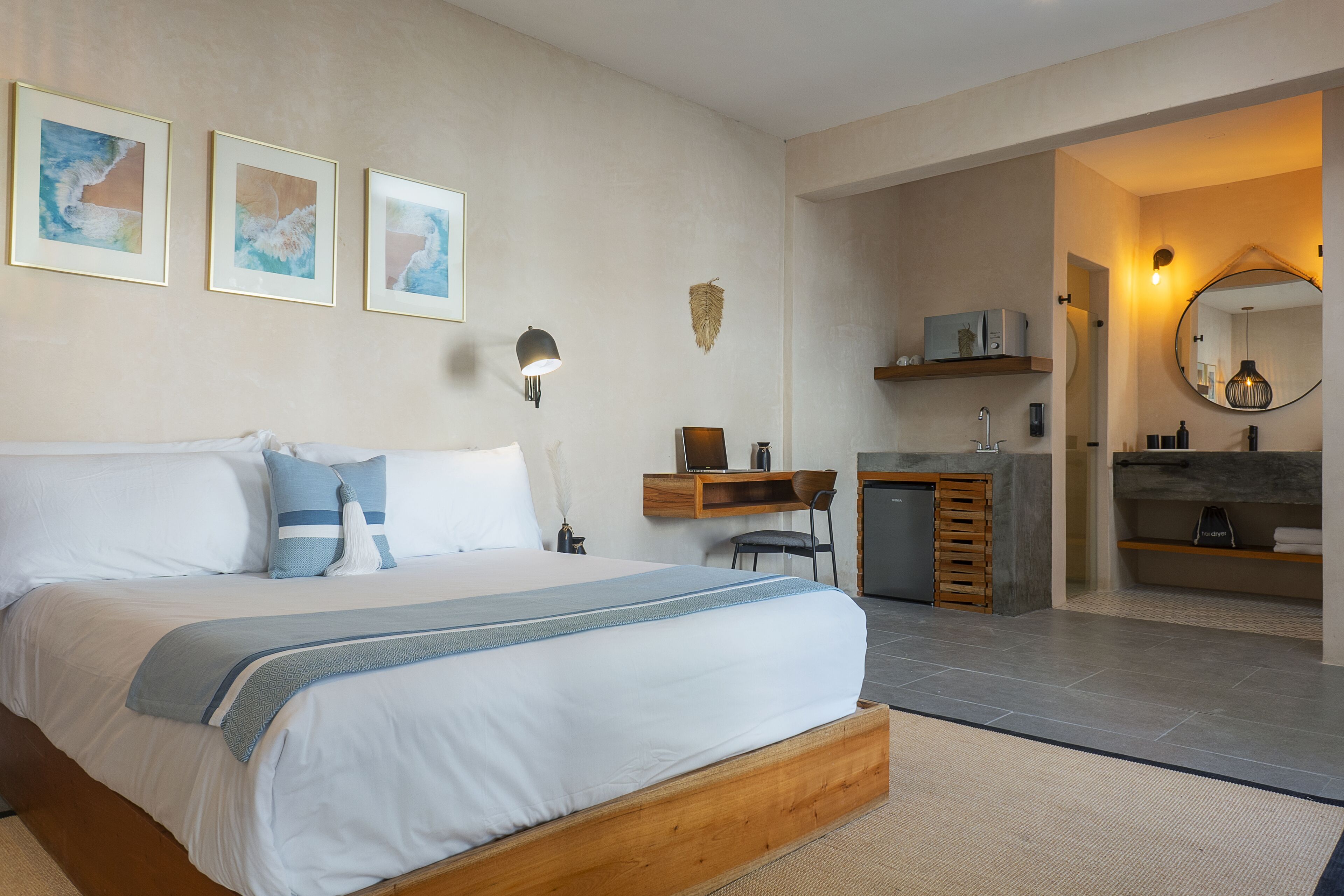 Un dormitorio con cama, mesita de noche y escritorio con silla. Hay un baño con lavamanos, espejo y estanterías.