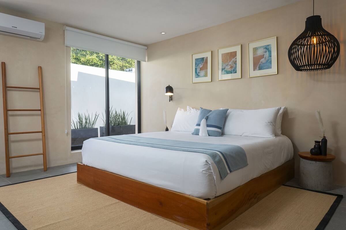 A bedroom with a bed, a ladder, a bedside table, a lamp, and artwork on the wall.