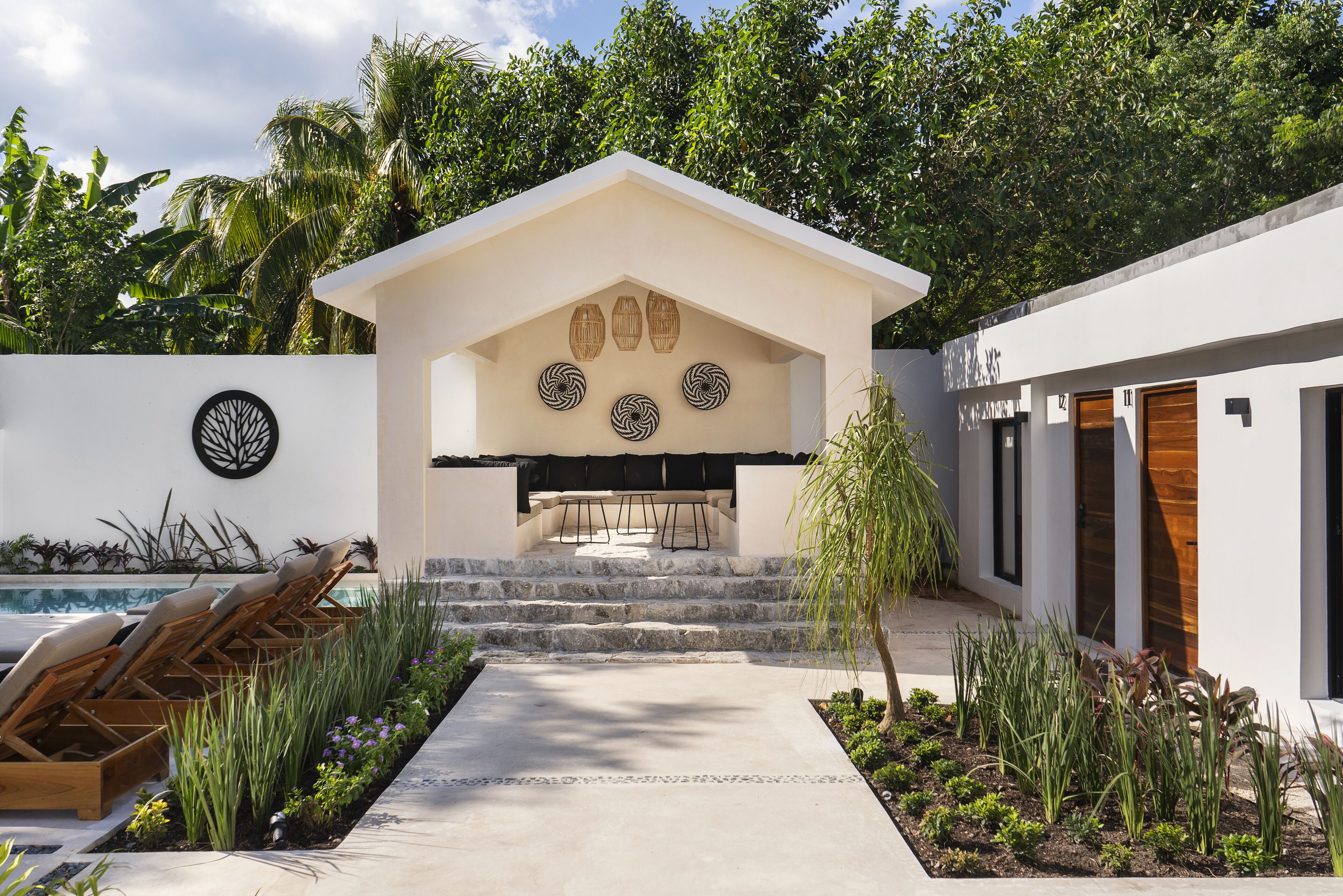 A modern outdoor seating area with a pool, surrounded by lush greenery and a white building with a unique wall design.