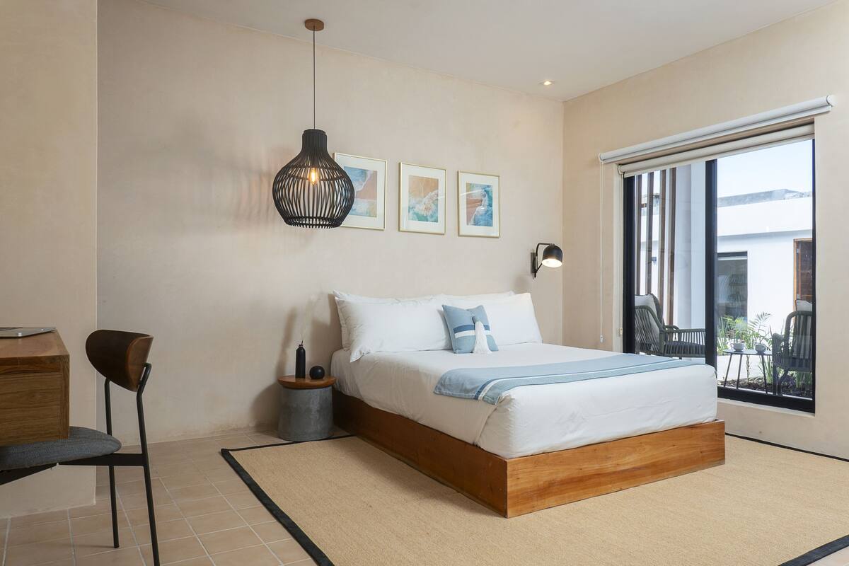 A bedroom with a wooden bed, a desk, a chair, a large window, and artwork on the wall.