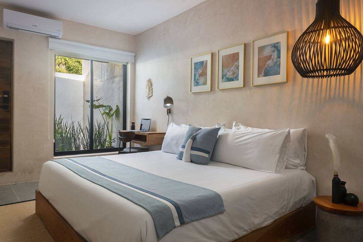 A bedroom with a large bed, a desk, and a window with a view of greenery.