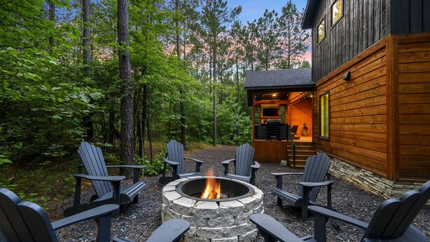 A cabin with a stone fire pit surrounded by Adirondack chairs, set in a forested area. - Pet-Friendly Cabin 5 minutes from town with stunning decor and views! (Broken Bow)