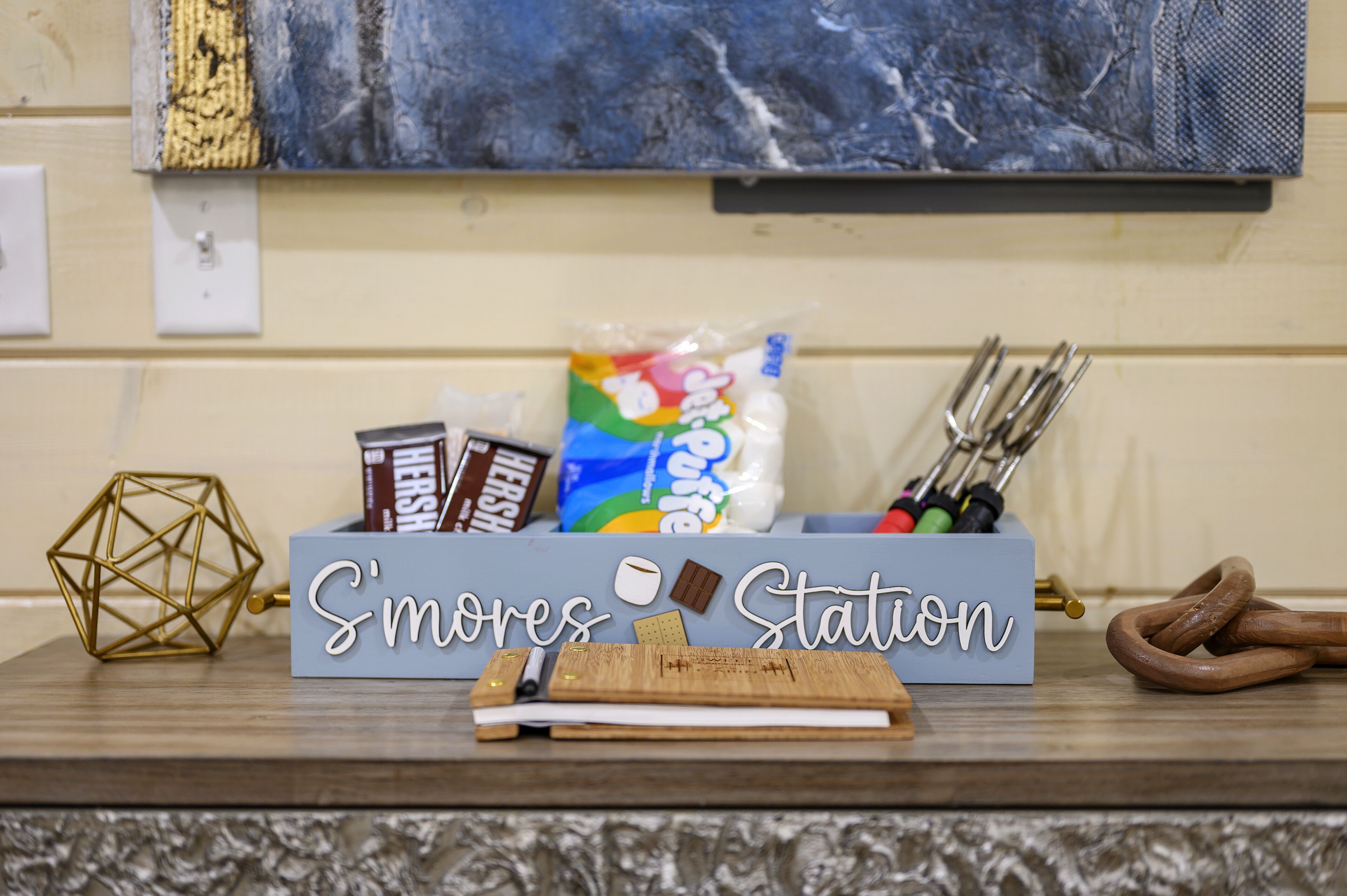 A 'S'mores Station' with a chocolate bar, marshmallows, and a s'mores kit on a wooden table.