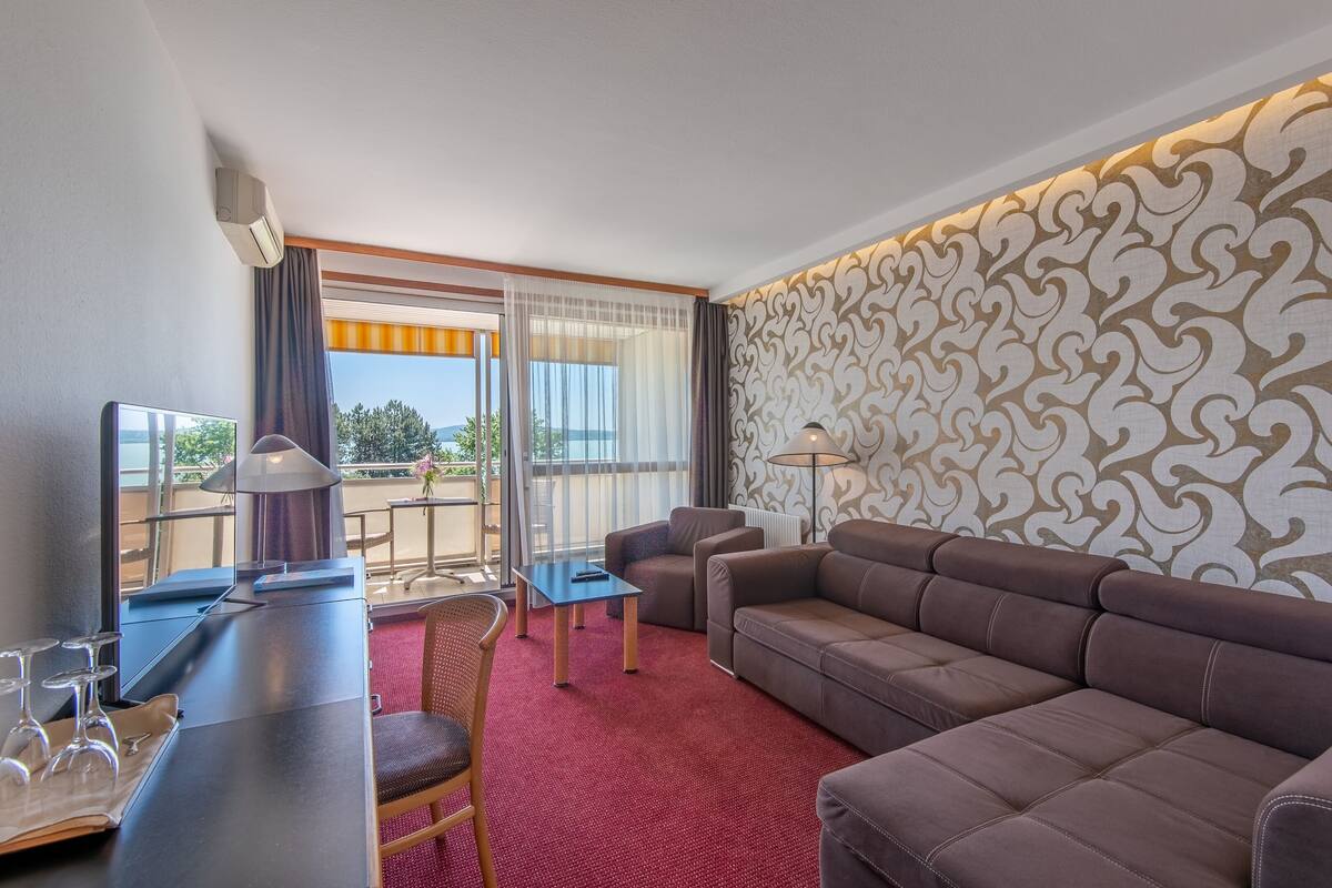 A hotel room with a sofa, a dining table, a chair, a TV, and a window with curtains.