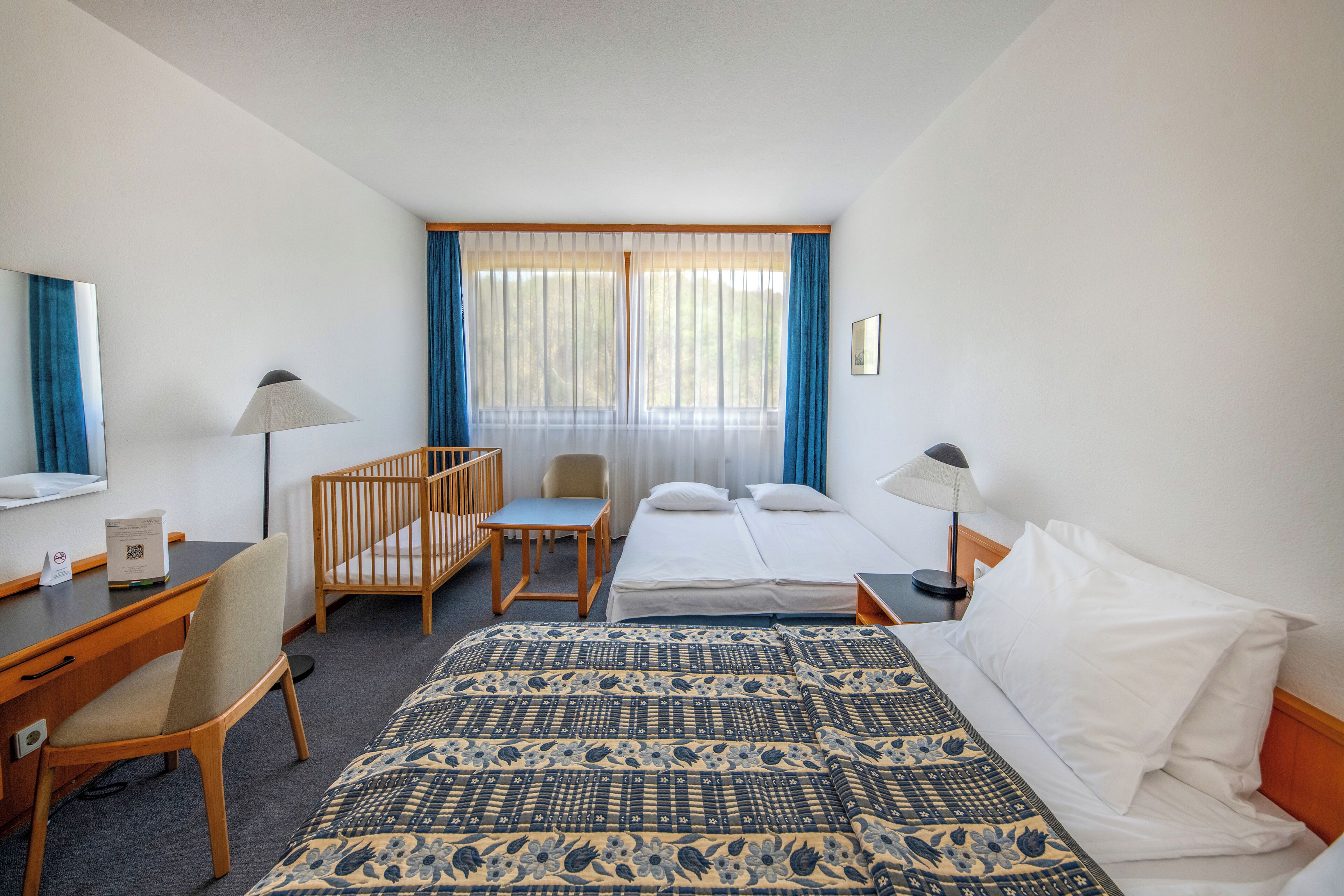 A hotel room with a bed, two bedside tables, a desk with a chair, a small table with a chair, and a crib.