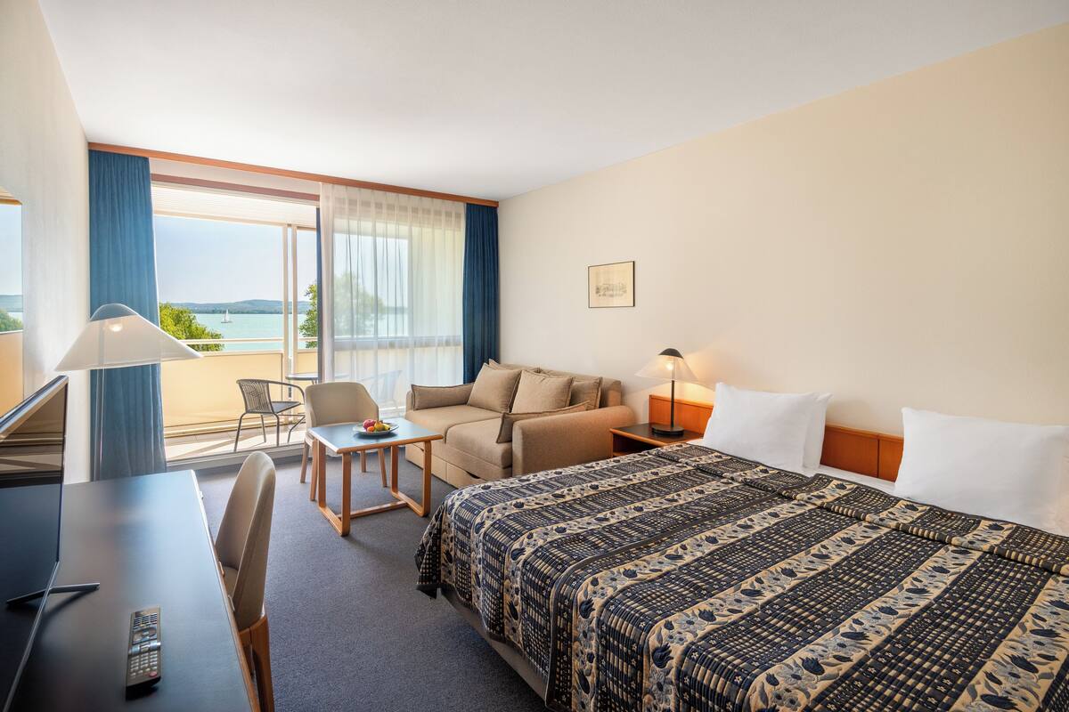 A hotel room with a bed, a desk with a remote control, a chair, a small table with a fruit bowl, a sofa, and a view of the sea through large windows.