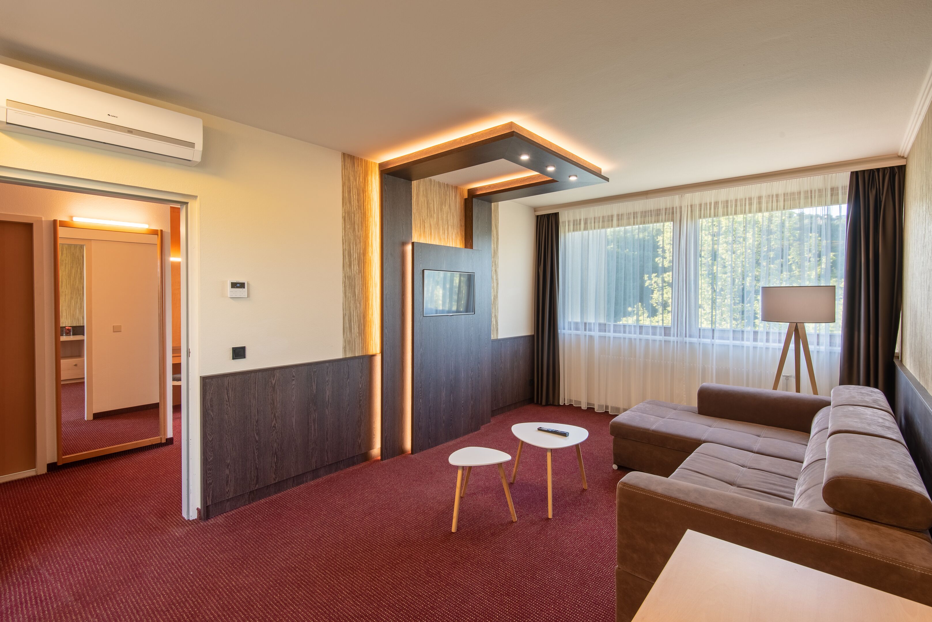 A hotel room with a red carpet, a sofa, two small tables, a lamp, and a large window with curtains.