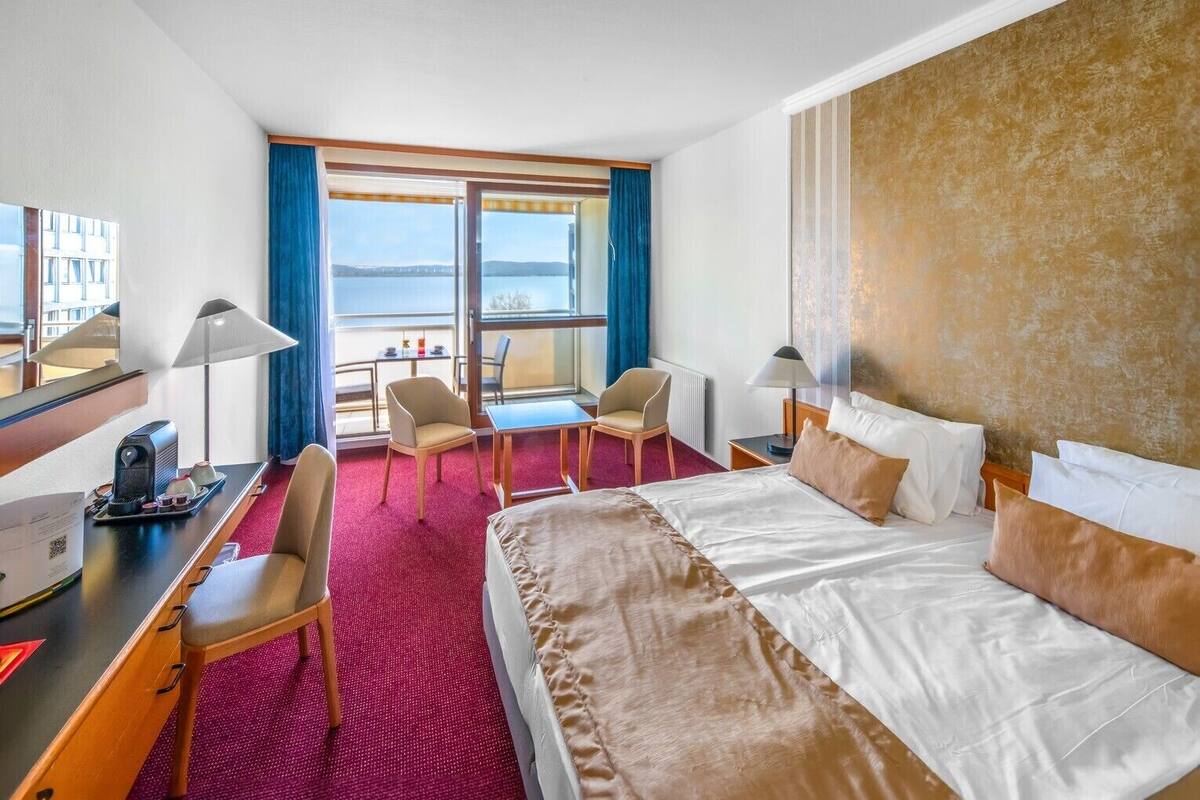 A hotel room with a bed, desk, chairs, and a view of the outdoors.