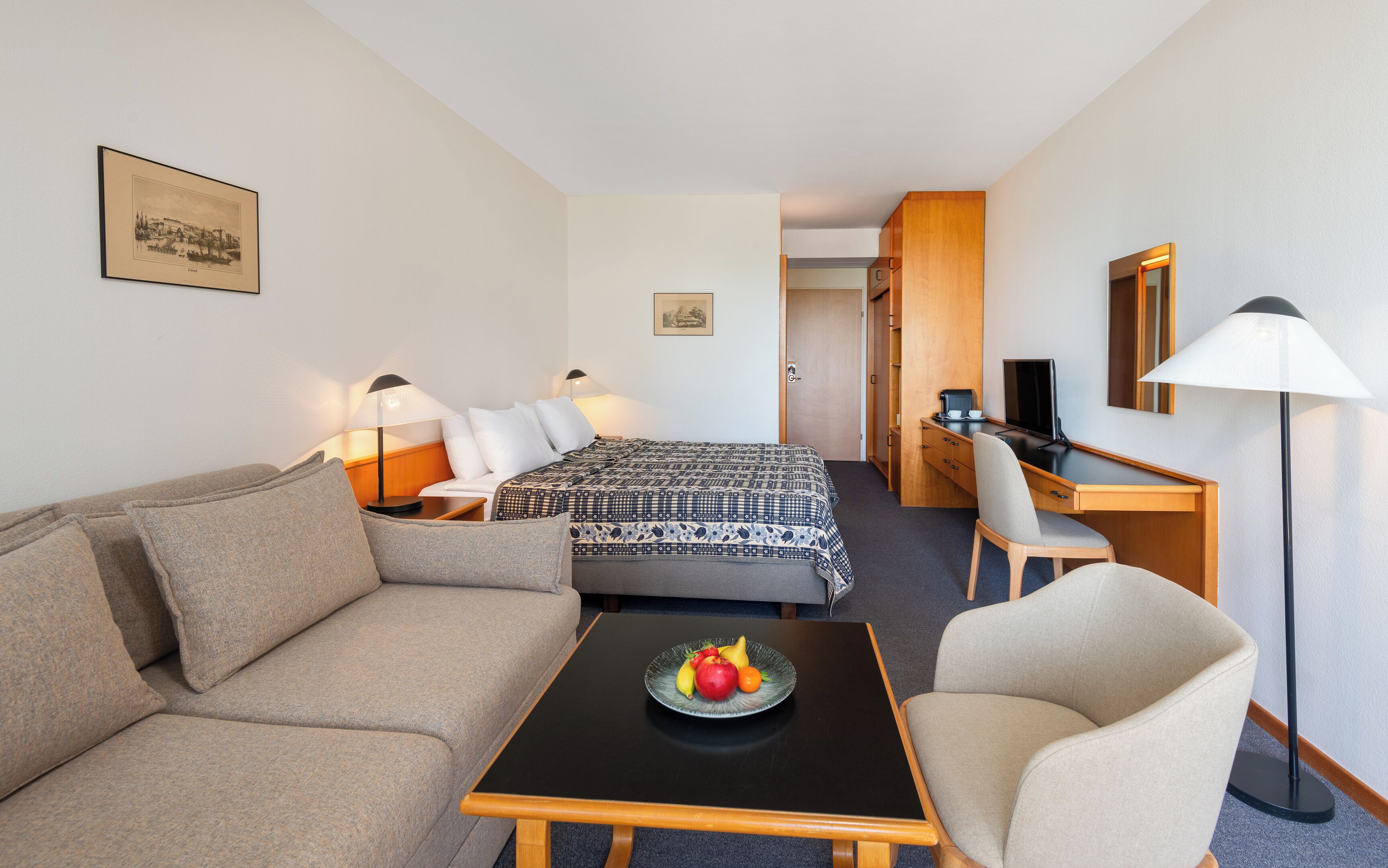 Habitación de hotel con cama, sofá, escritorio, silla, una mesita con fruta y lámpara.