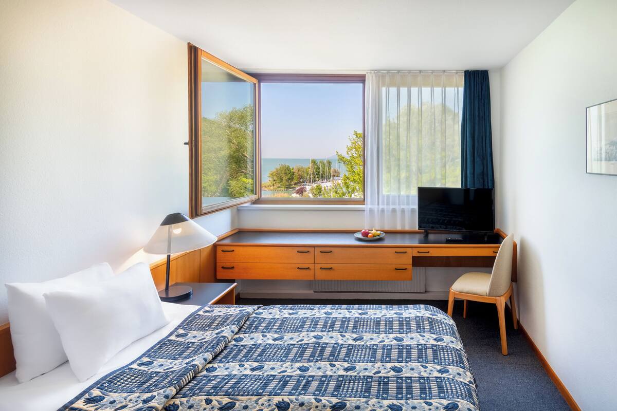 A hotel room with a bed, a desk with a computer, a chair, a TV, and a window with a view of the sea.