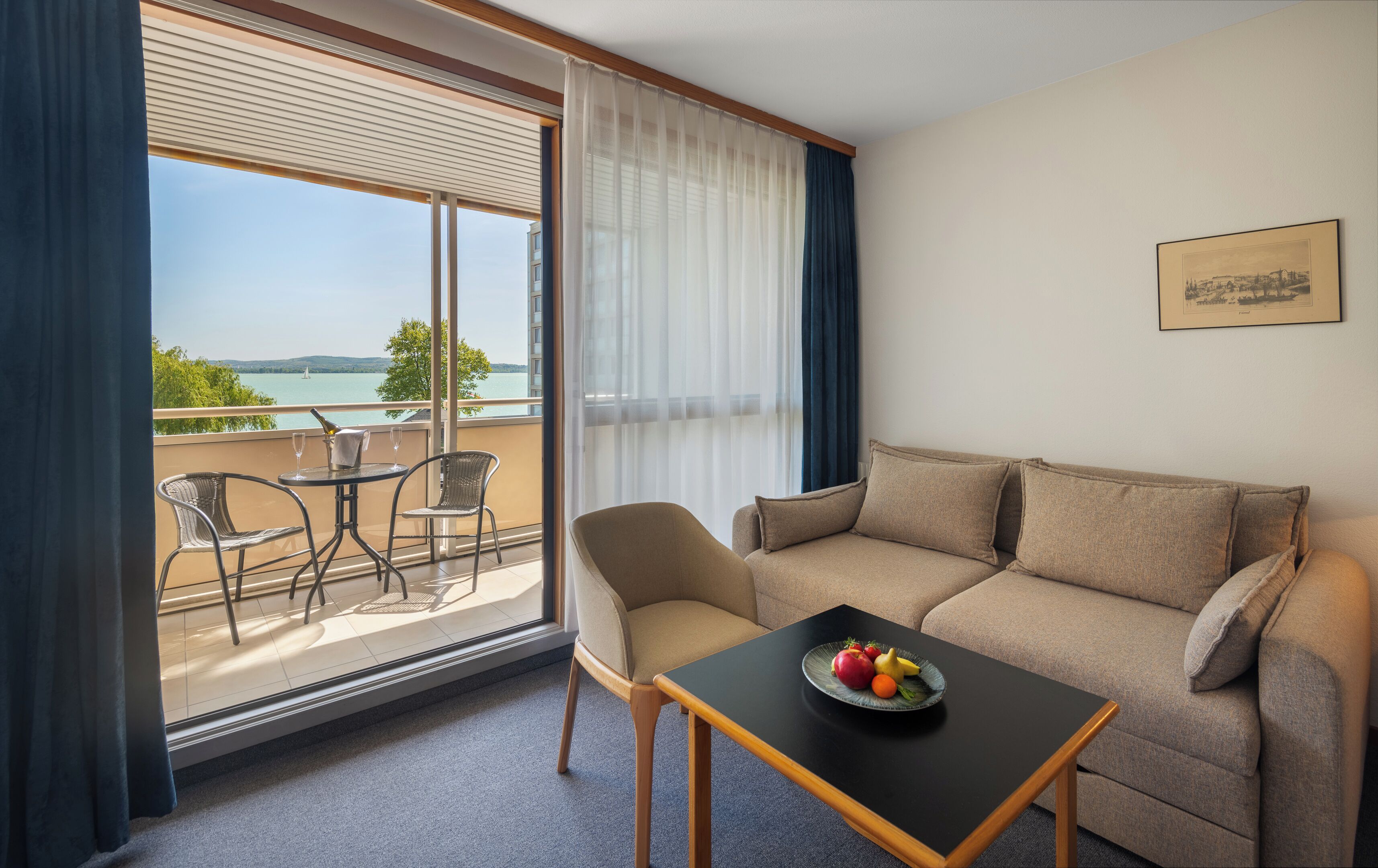 A hotel room with a balcony, a sofa, a table with fruit, and a painting on the wall.