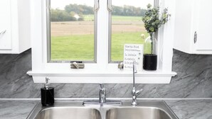 Een keuken met een dubbele spoelbak, een raam met uitzicht op een groen veld en een bord met een lijst met stappen.