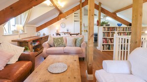 A cozy loft-style living room with exposed wooden beams, a wooden dining table, a sofa, and a bookshelf. - The Old Barn (Colyton)