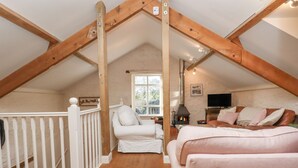 A cozy attic bedroom with a sloped ceiling, wooden beams, a white railing, a bed, a sofa, a fireplace, and a TV. - The Old Barn (Colyton)