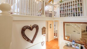 A spacious loft with a high ceiling, exposed wooden beams, a white railing, and a large bookshelf filled with books. - The Old Barn (Colyton)
