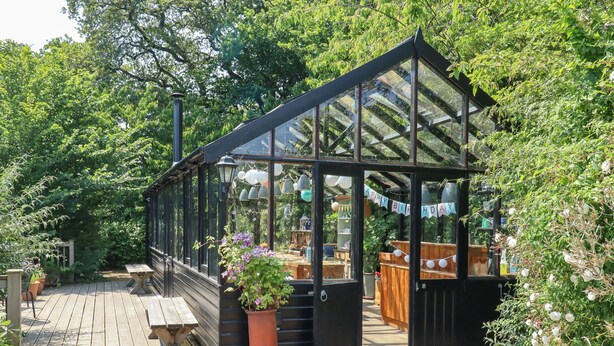 A greenhouse with a wooden deck, a table, and potted plants. - The Old Barn (Colyton)