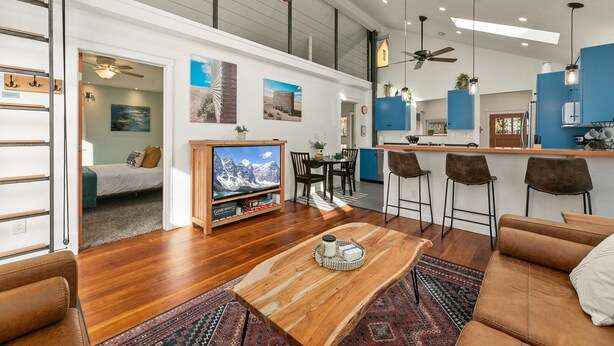 A spacious living area with a wooden coffee table, a TV, and a sofa. A kitchen with blue cabinets and a bar area is visible in the background. - Vibrant Downtown Bungalow – Walk Everywhere! (Fort Collins)