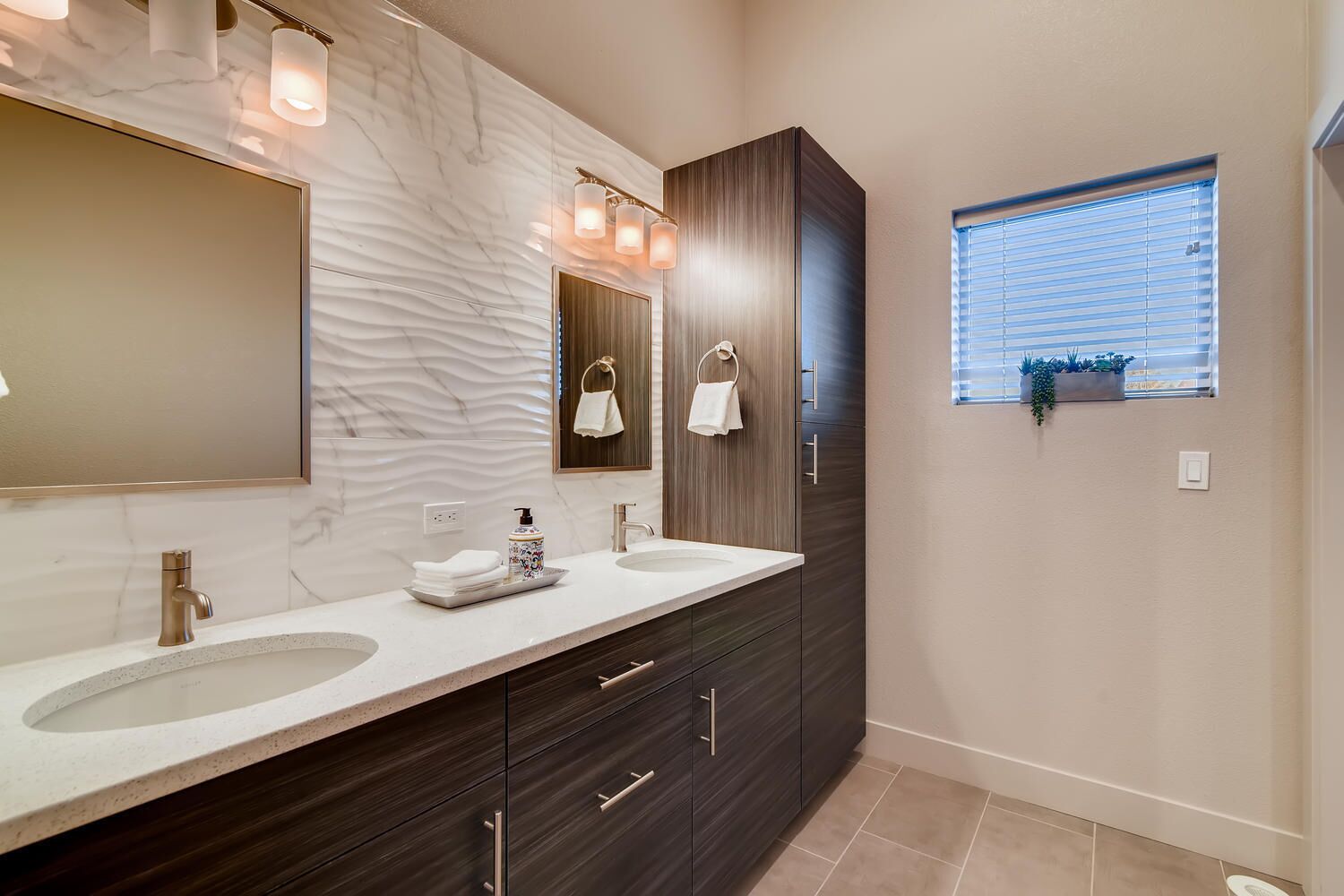 A modern bathroom with two sinks, dark wood cabinets, a marble backsplash, and a window with plants.