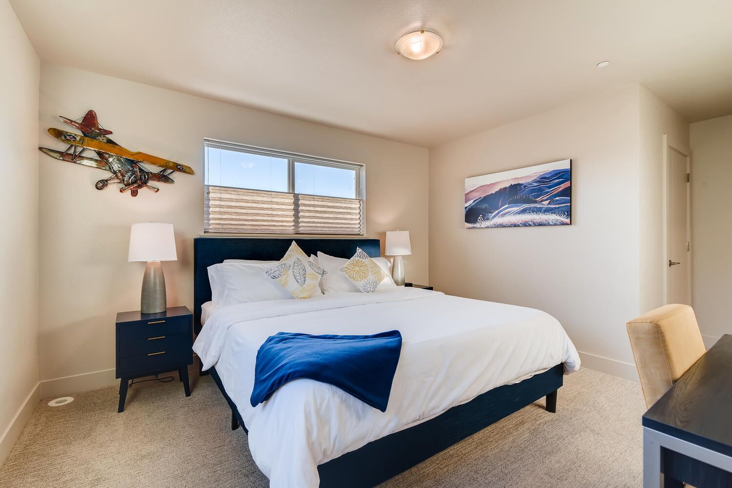 A bedroom with a large bed, two bedside tables with lamps, a window with blinds, a wall-mounted artwork, and a door.