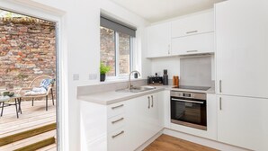 Une cuisine moderne avec des meubles blancs, un four encastré et une fenêtre donnant sur un mur en pierre.