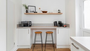 Une cuisine compacte équipée d’un grille-pain encastré, d’une machine à café et d’une étagère ornée d’une plante et d’un tableau. On y trouve également deux tabourets et un comptoir blanc.