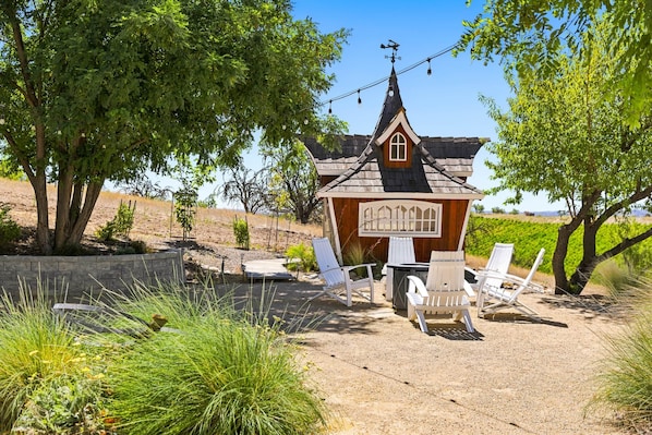 A rustic wooden structure with a pitched roof and a small window, surrounded by white chairs and a table, set in a landscaped garden with trees and a clear sky.