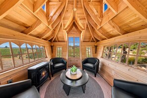 Interior de madera con chimenea, dos sillones de cuero, una mesa redonda con una planta y ventanas amplias que ofrecen vistas al exterior.