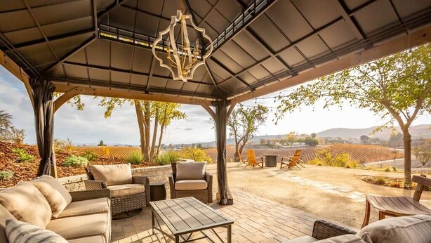 Un área de estar al aire libre techada con mobiliario de mimbre, una mesa y vistas a viñedos.