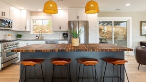 Cocina moderna con gabinetes blancos, una isla de madera con taburetes, un refrigerador de acero inoxidable y lámparas colgantes de mimbre.
