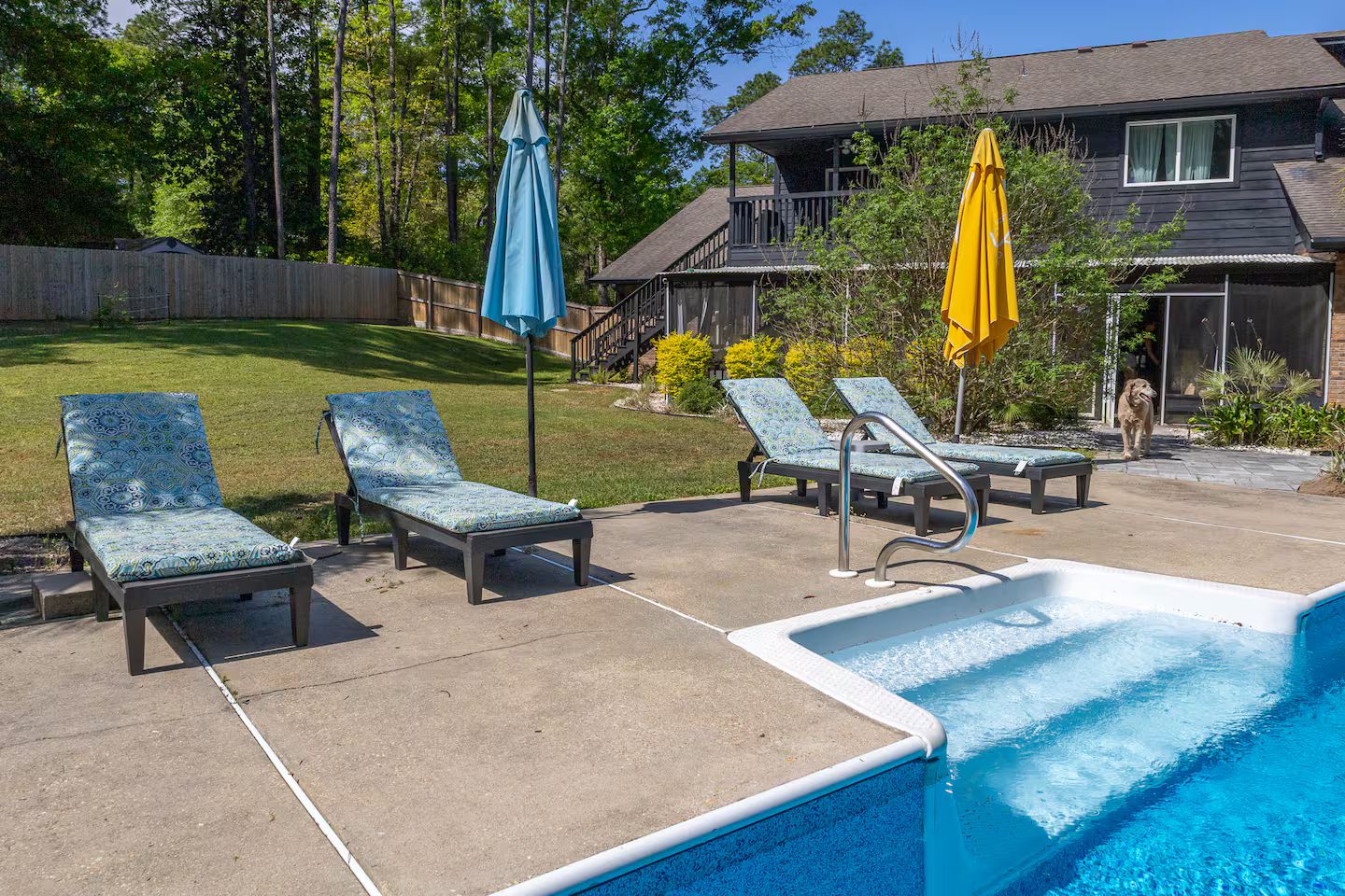A swimming pool with lounge chairs and umbrellas, a wooden deck, and a house in the background.