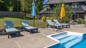 A swimming pool with lounge chairs and umbrellas, a wooden deck, and a house in the background.