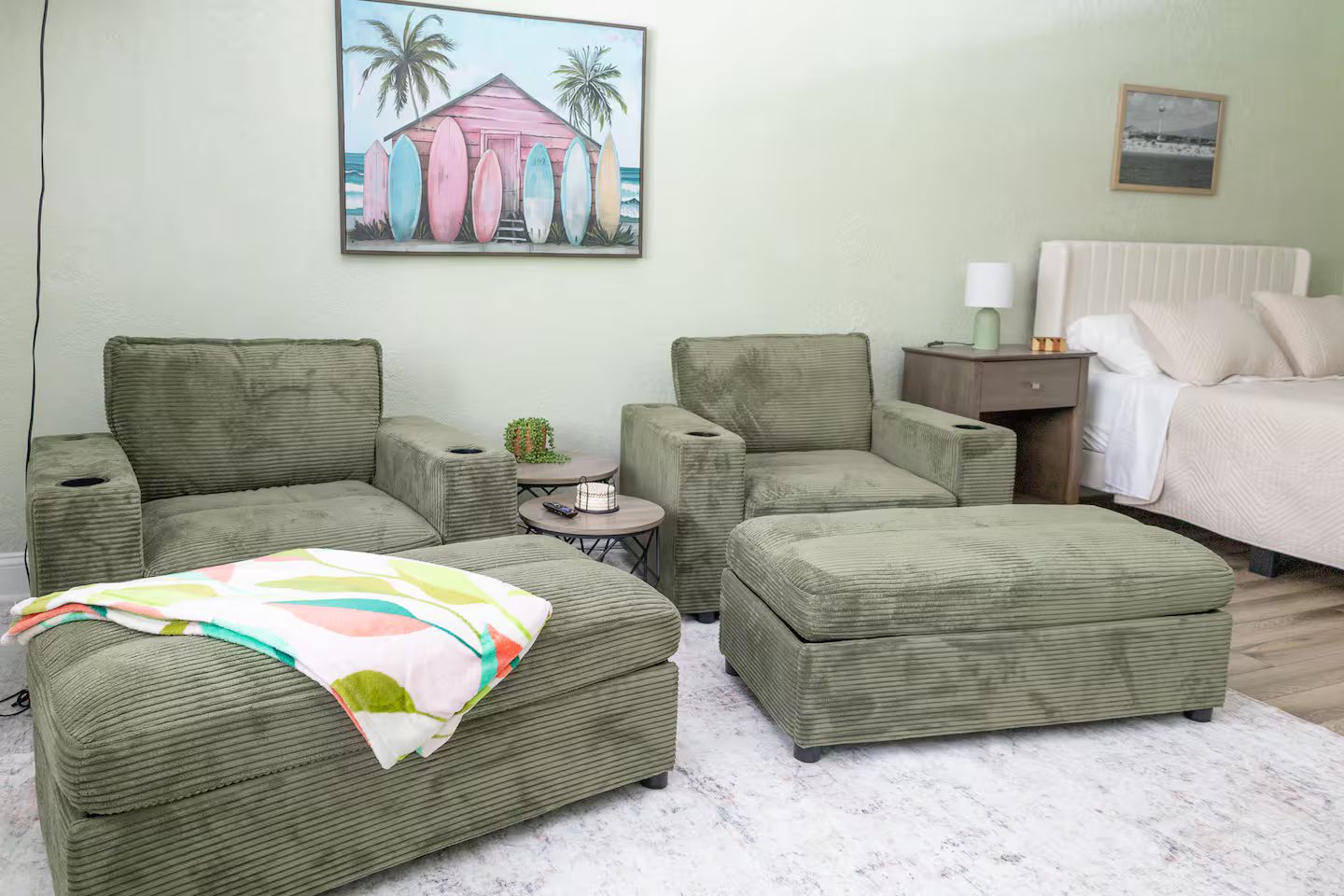Ein Zimmer mit einem Bett, zwei grünen Sesseln, einem kleinen Tisch mit einer Pflanze und einer farbenfrohen Decke auf einem Hocker.