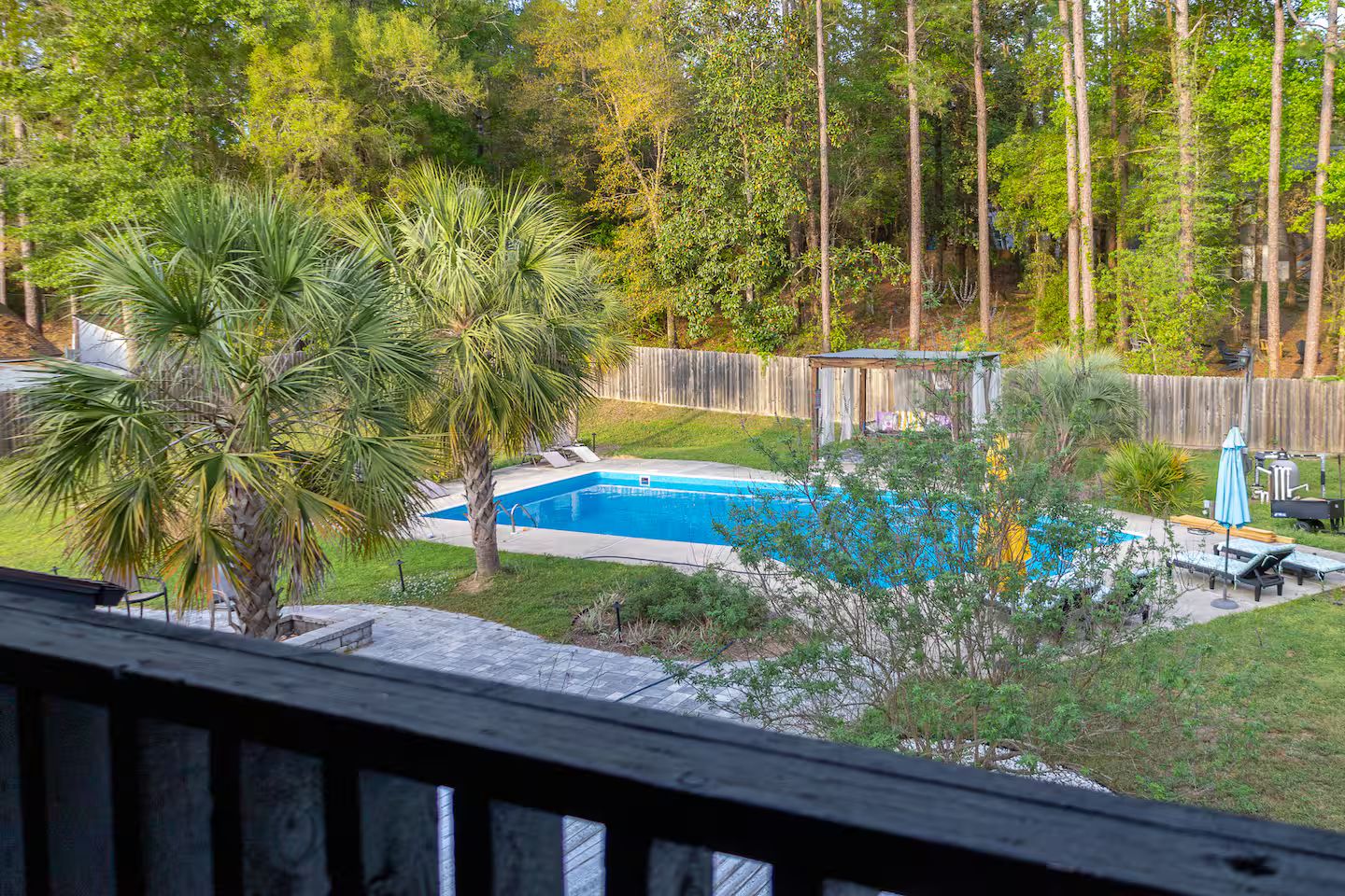Ein Garten mit Swimmingpool, einer Holzterrasse und einem von Bäumen gesäumten, eingezäunten Bereich.