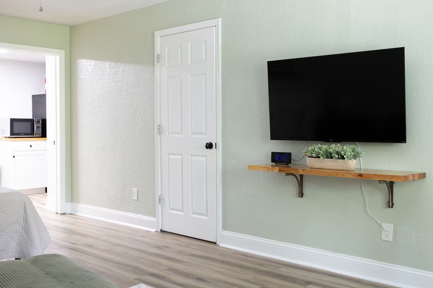 A room with a white door, a mounted TV, a shelf with a plant, and a microwave in the background.