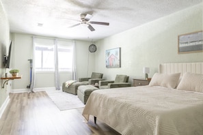 A bedroom with a bed, two chairs, a desk, a ceiling fan, and a window.