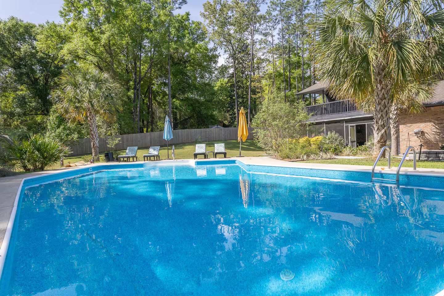 A rectangular swimming pool with a clear blue surface, surrounded by trees and a wooden deck with lounge chairs.