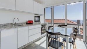 A modern kitchen with white cabinets, a granite countertop, and a red toaster. There is a dining area with a glass table and chairs, and large windows offering a view of the city. - Villa Winter (Funchal)