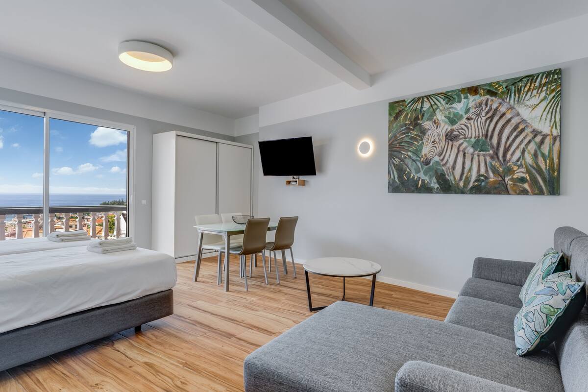 A modern bedroom with a large bed, a grey sofa, a dining area, and a TV mounted on the wall.