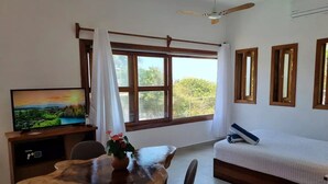 Ein Zimmer mit einem Holzesstisch, zwei Stühlen, einem Fernseher, einem Bett mit weißer Bettdecke und einem Fenster mit Vorhängen.
