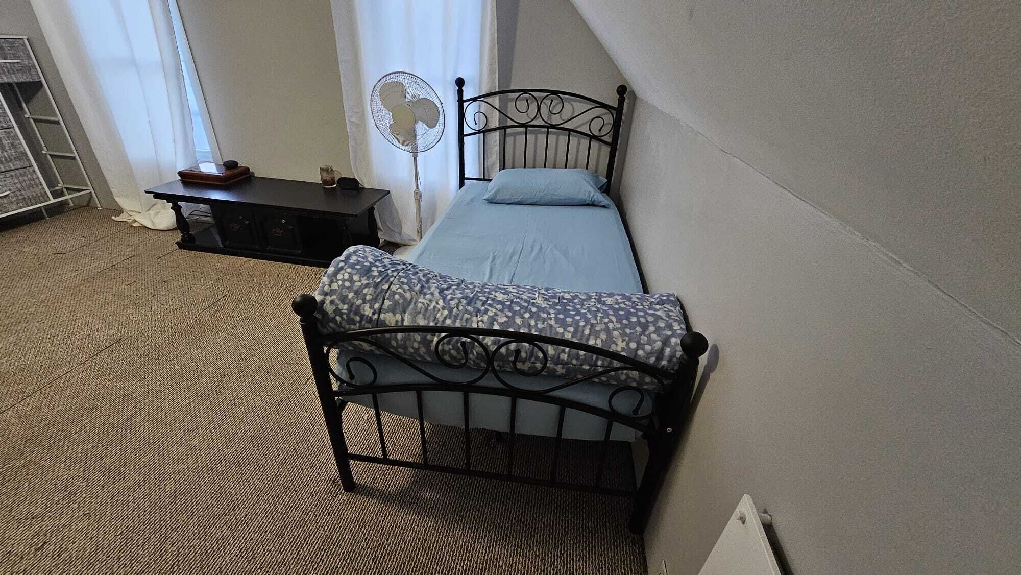Une petite chambre à coucher, bien rangée, avec un lit simple, une table de chevet, un ventilateur sur pied et une fenêtre avec des rideaux.
